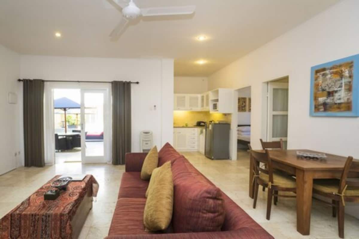 A living room with a sofa, a dining table with chairs, a kitchen in the background, and a door leading to an outdoor area.