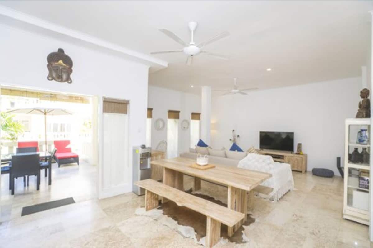 A modern living room with a wooden dining table, a sofa, a TV, and ceiling fans.