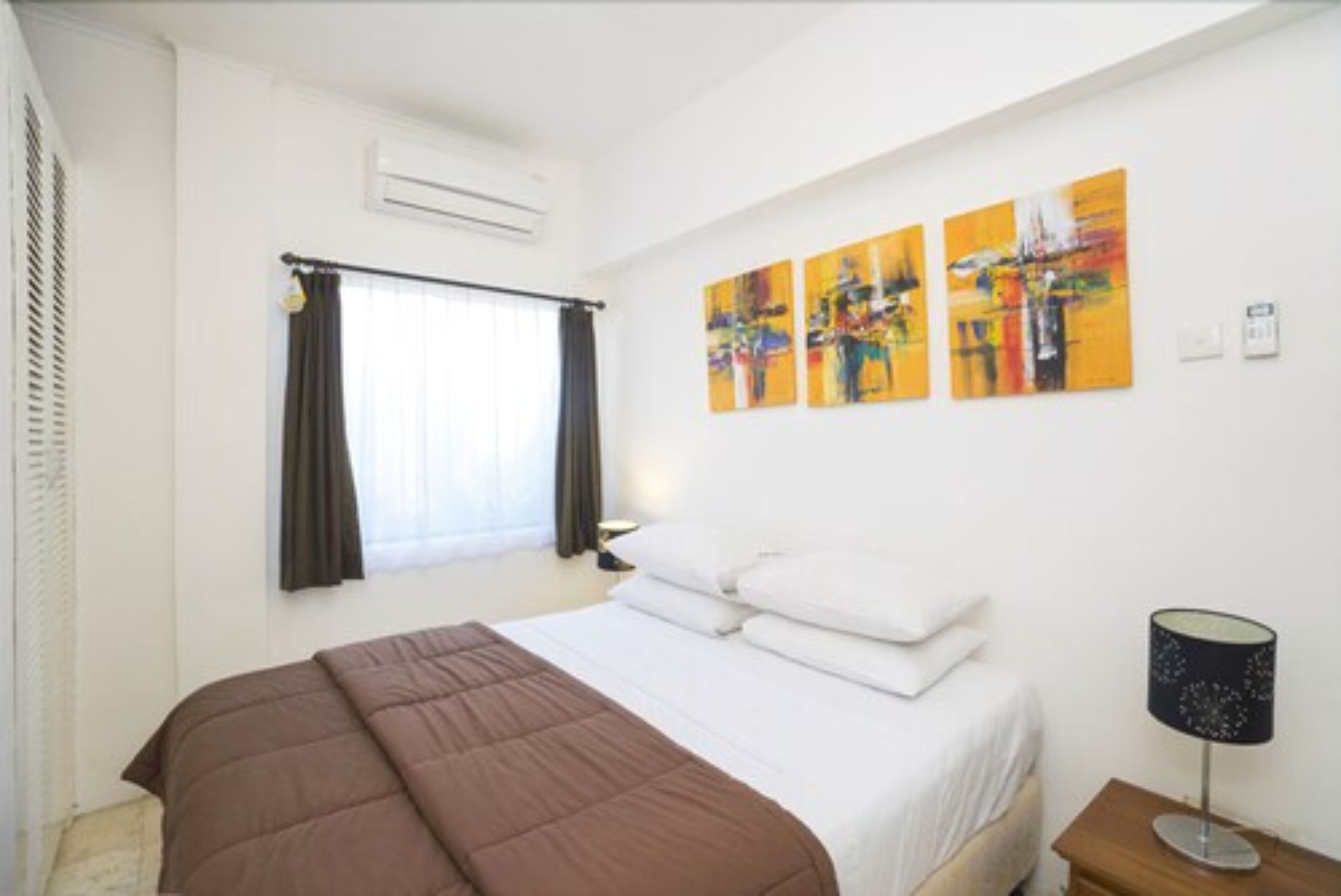 A hotel room with a bed, a window with curtains, a wall-mounted air conditioner, and three abstract paintings.