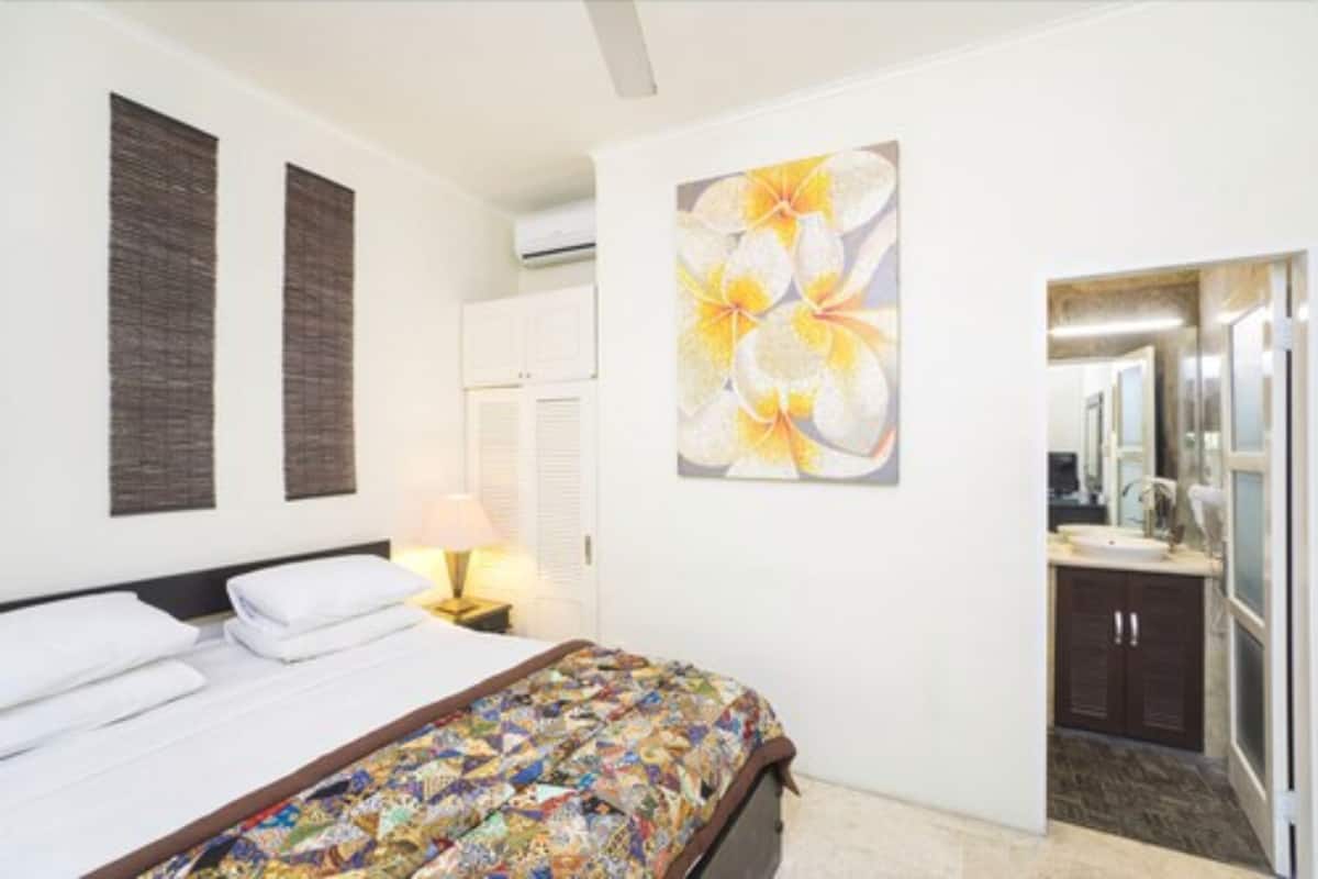 A hotel room with a bed, a nightstand, a painting, a bathroom with a mirror, and a cabinet.