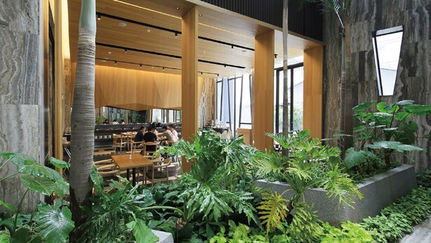 Un espacio interior moderno con mesas y sillas de madera, rodeado de abundante follaje y grandes ventanales que permiten la entrada de luz natural.