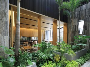 Un espacio interior moderno con mesas y sillas de madera, rodeado de abundante follaje y grandes ventanales que permiten la entrada de luz natural.