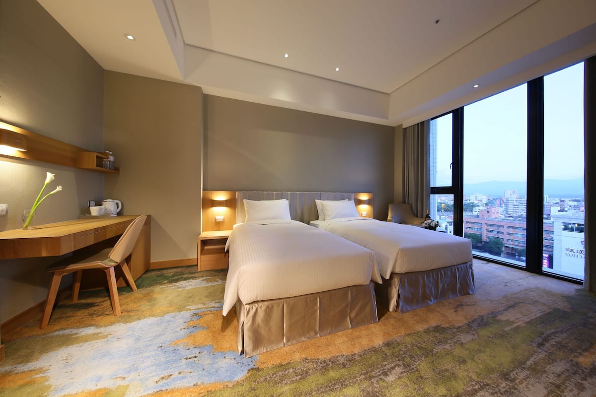 Habitación de hotel con dos camas, un escritorio, una silla y un ventanal con vistas a la ciudad.