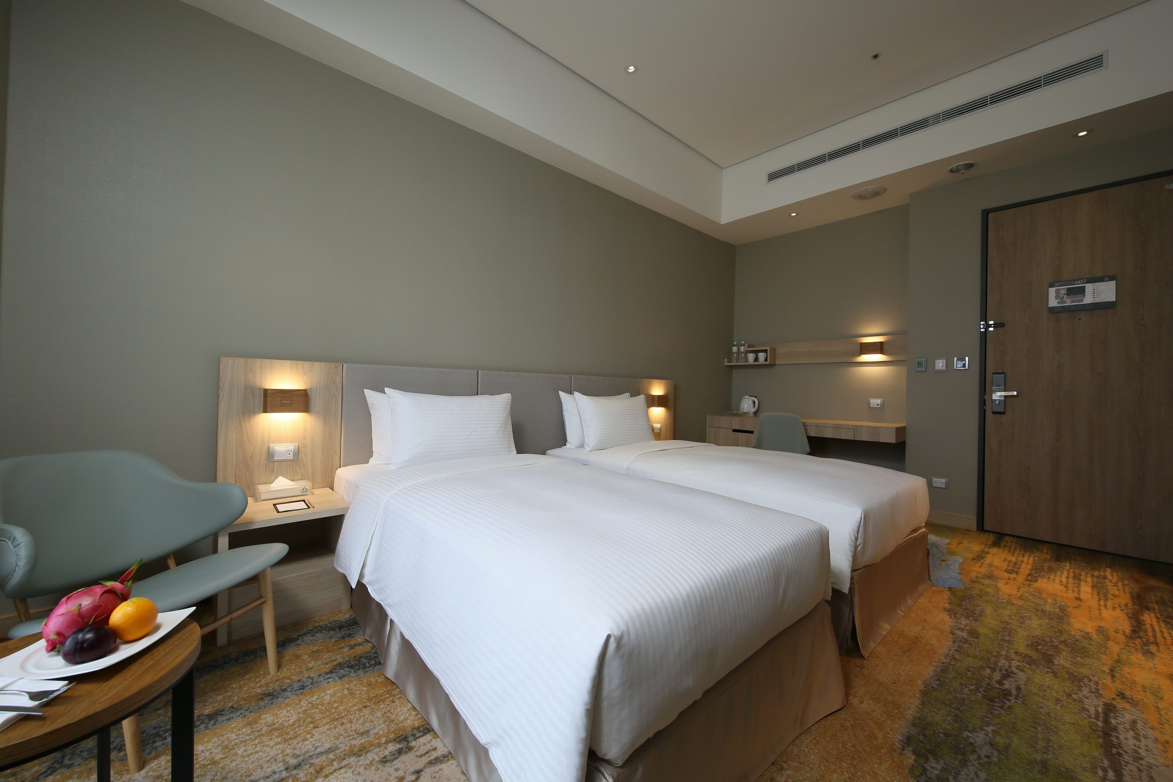 A hotel room with two beds, a desk, a chair, and a small table with fruit.