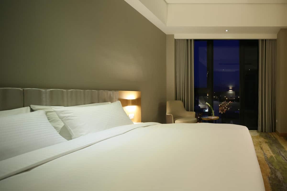 Habitación de hotel con una cama grande, una mesita de noche con lámpara, una silla y vistas a la ciudad a través de un ventanal amplio.