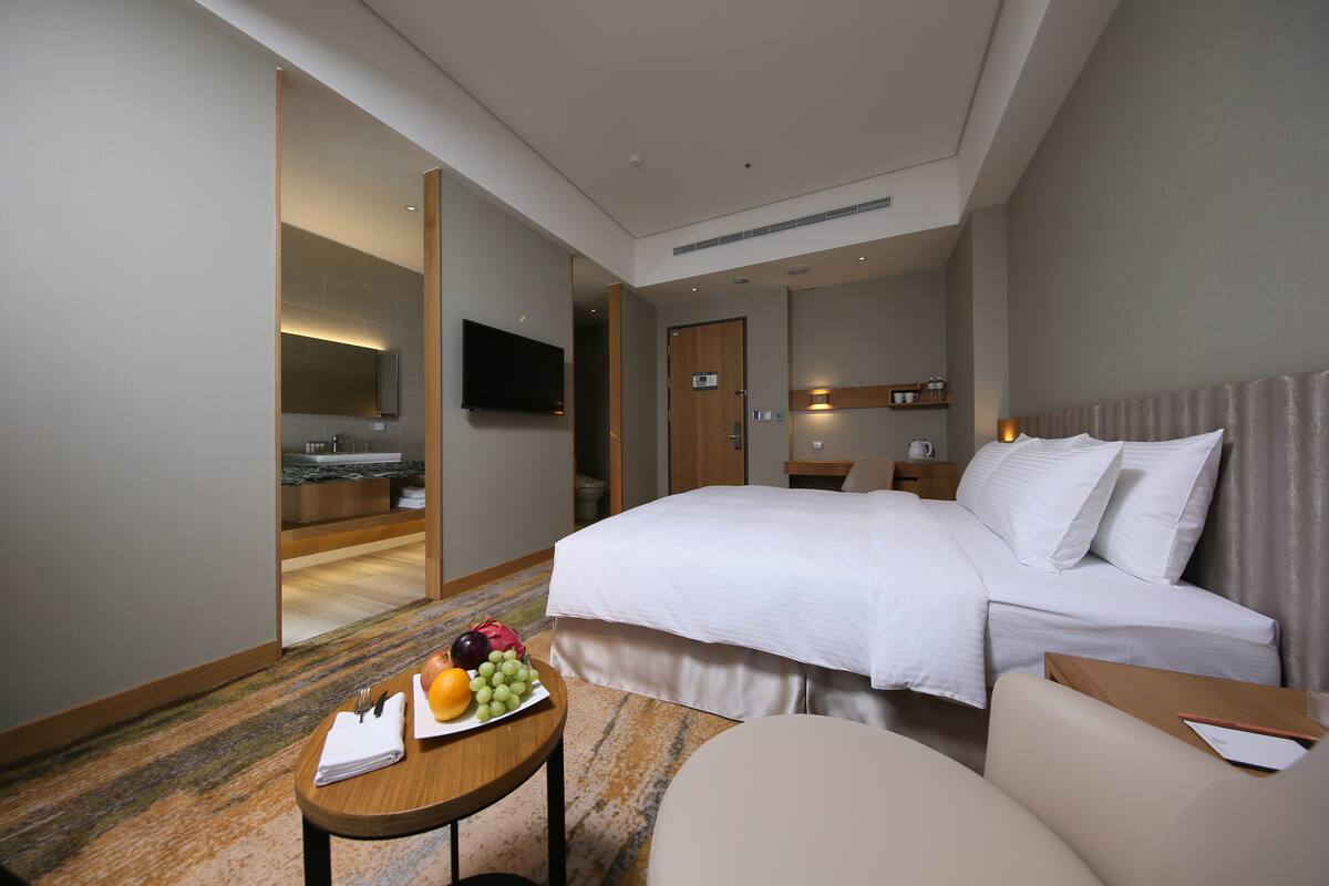 Habitación de hotel con una cama grande, una mesita con fruta, una silla, un televisor y un baño visible a través de una puerta abierta.