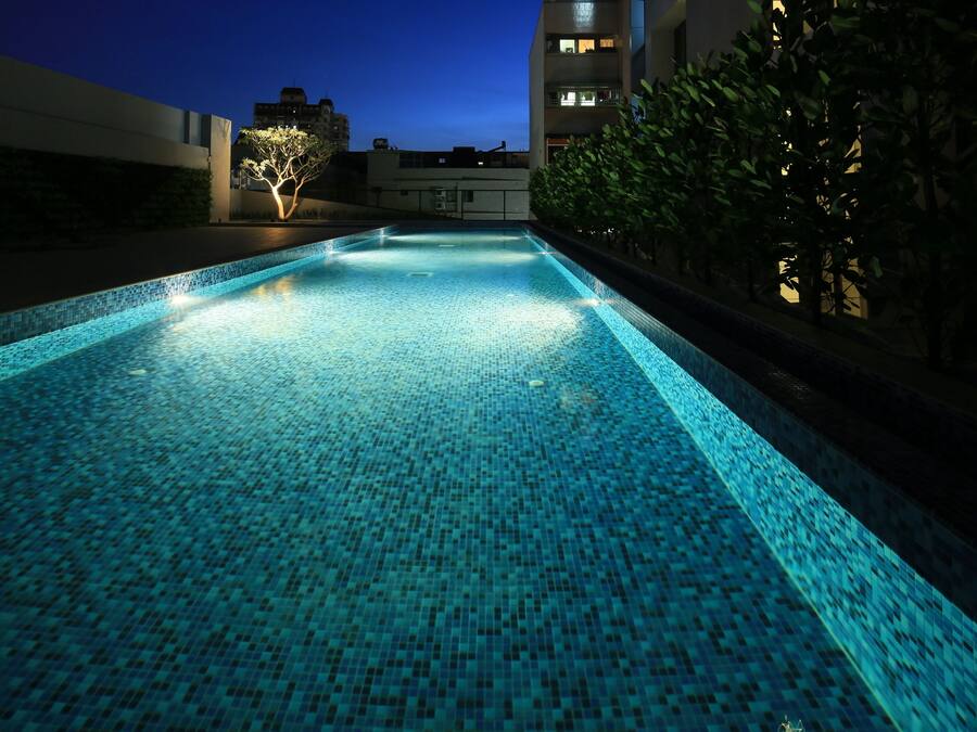 A swimming pool with blue mosaic tiles, surrounded by a low wall and greenery, with buildings in the background.