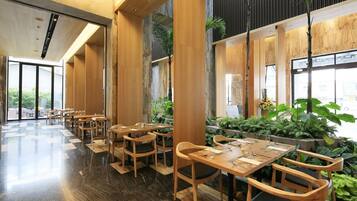 Un restaurante moderno con mesas y sillas de madera, ventanas amplias y plantas de interior.