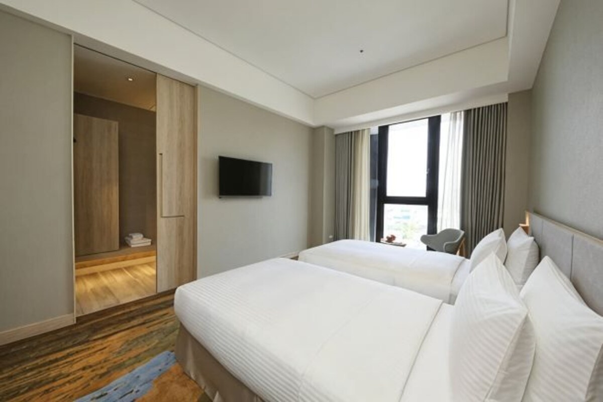 Quarto de hotel com duas camas, televisão, janela com cortinas e chão de madeira.