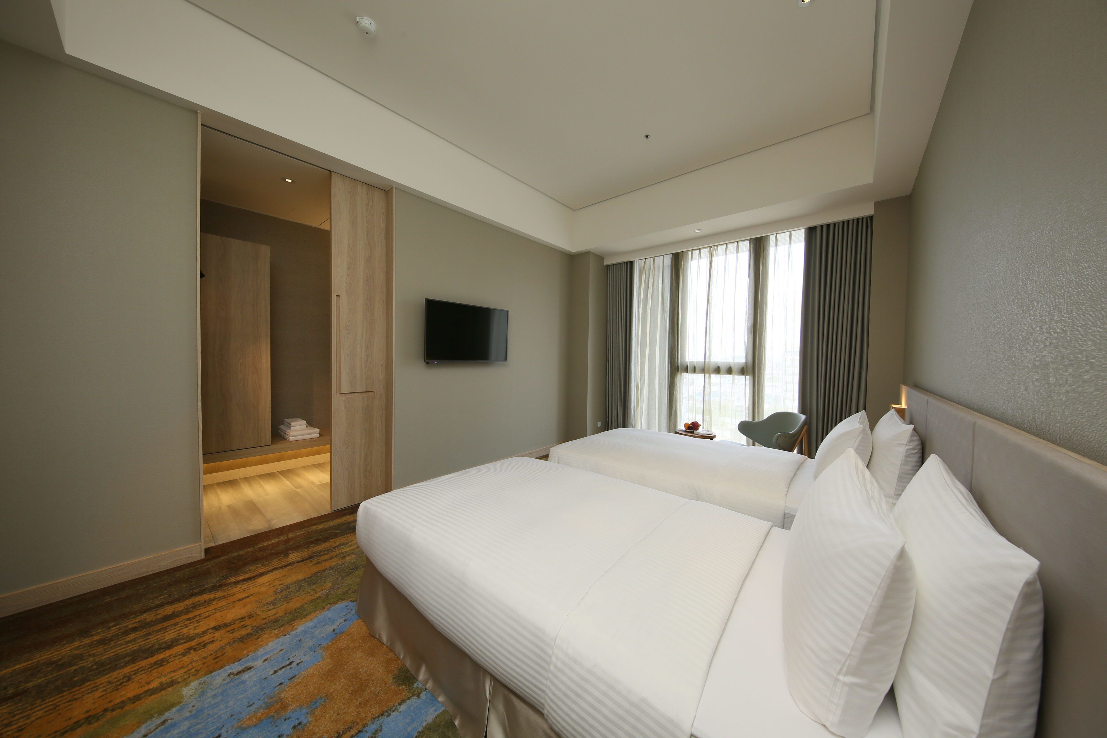 A hotel room with two beds, a TV, a large window with curtains, and a wooden wardrobe.