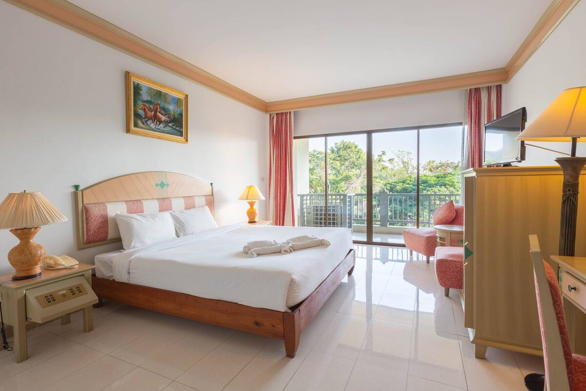Habitación de hotel con cama, lámparas de noche, televisor, escritorio y balcón con vistas a zonas verdes.