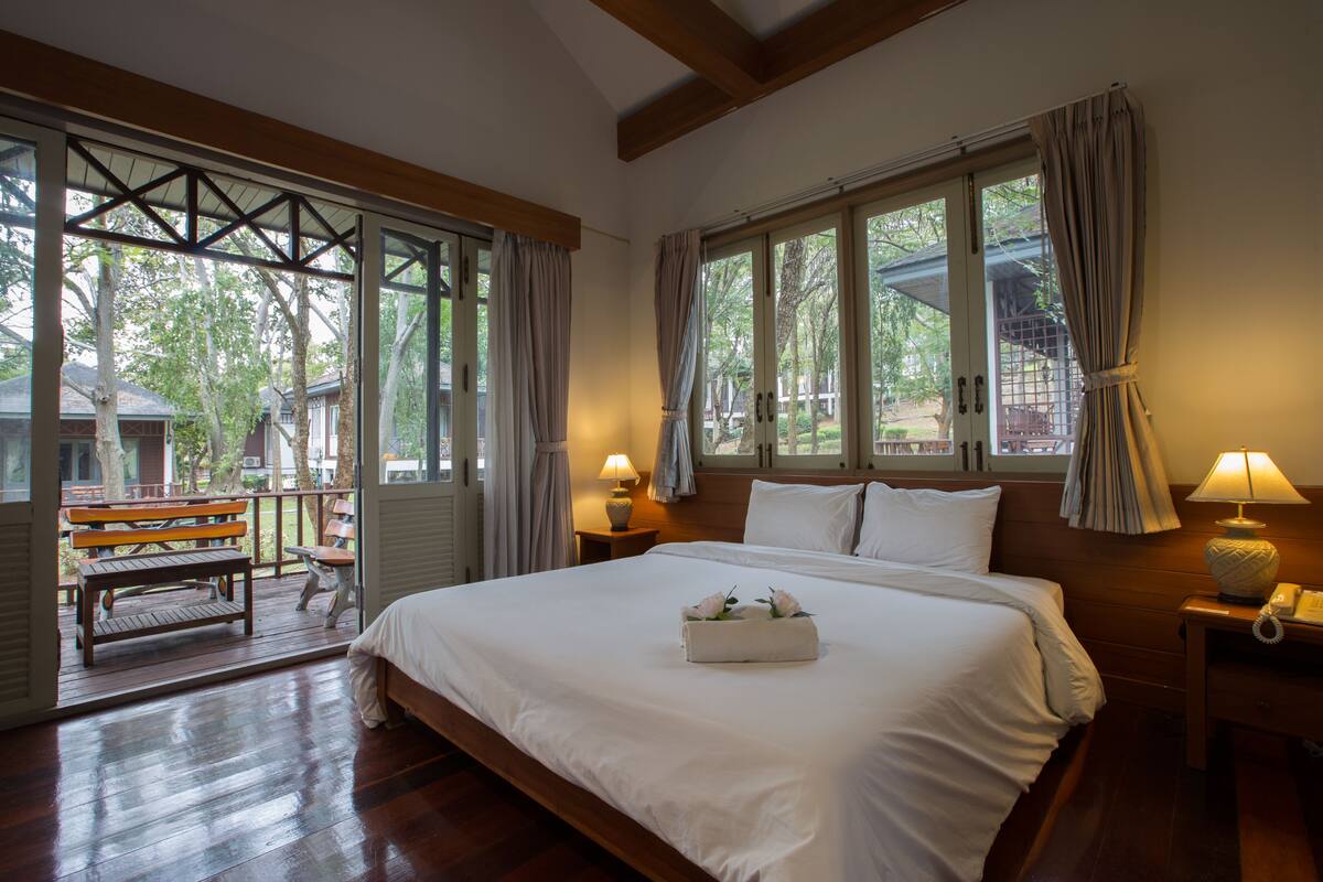 Un dormitorio con una cama grande, cabecera de madera y dos lámparas de noche. Se ve una ventana con cortinas y vistas al exterior.