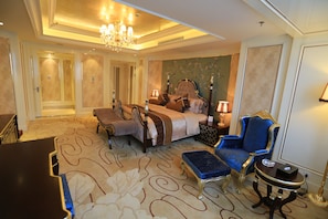 A spacious hotel room with a large bed, a sitting area with a blue armchair, a small table, and a chandelier.