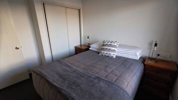 A neatly made bed with a grey quilt, a white pillow, and a patterned pillow on a wooden nightstand.