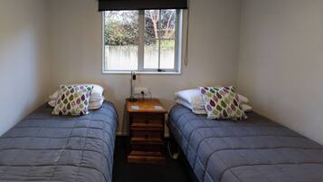 A room with two single beds, a small wooden nightstand, a window with a view of trees, and a wall-mounted light fixture.