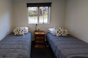 A room with two single beds, a small wooden nightstand, a window with a view of trees, and a wall-mounted light fixture.