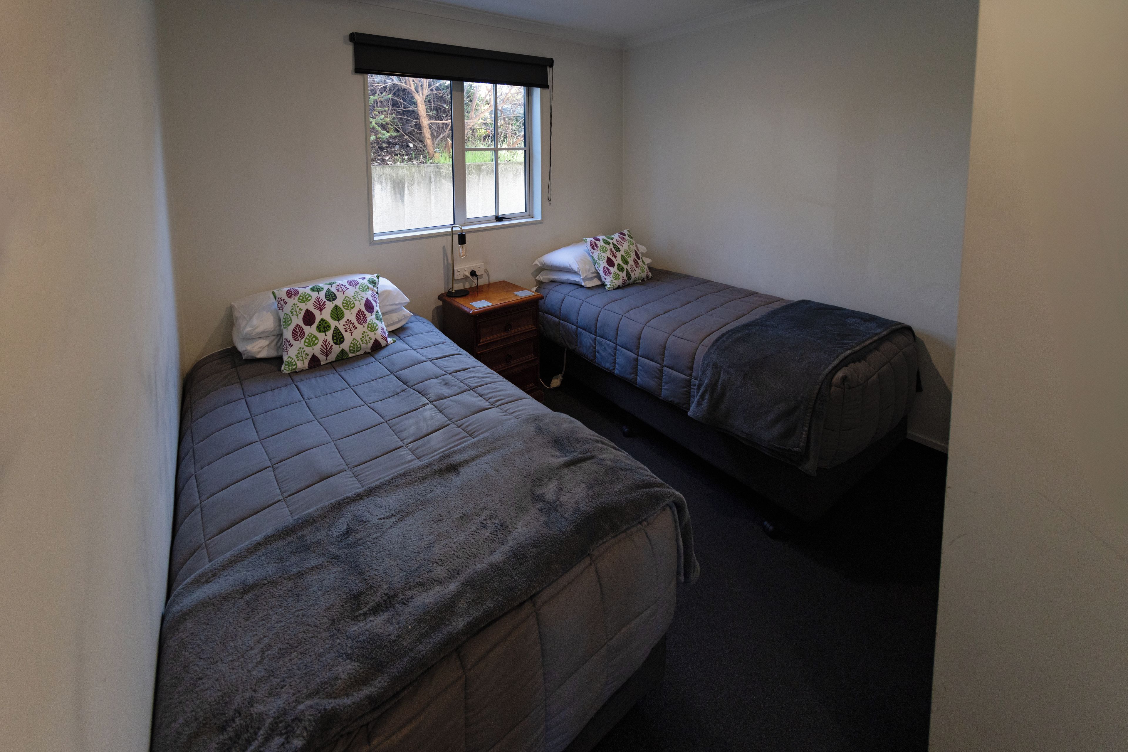 A small, single-room accommodation with two single beds, a nightstand, a window with a blind, and a view of the outdoors.