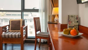 A hotel room with a wooden chair and sofa, a table with fruit, a lamp, and a view of buildings through large windows. - Bounty Hotel (Hanoi)