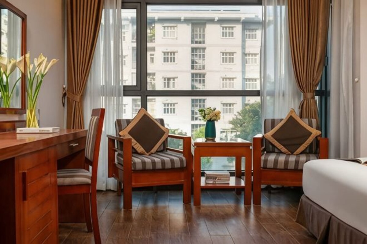 A hotel room with a wooden desk, two chairs with cushions, a small table with a vase, and a large window with curtains.
