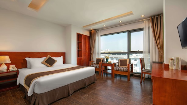 A hotel room with a large bed, a desk with a chair, a TV, and a view of the city through the window. - Bounty Hotel (Hanoi)