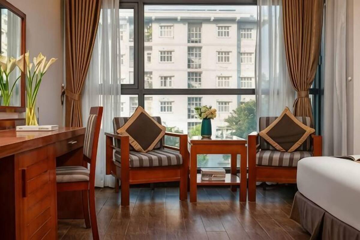 A hotel room with a wooden desk, two chairs with cushions, a small table with a vase, and a large window with curtains.