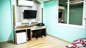 Una habitación con escritorio, televisor en la pared, refrigerador pequeño y ventana con vista.