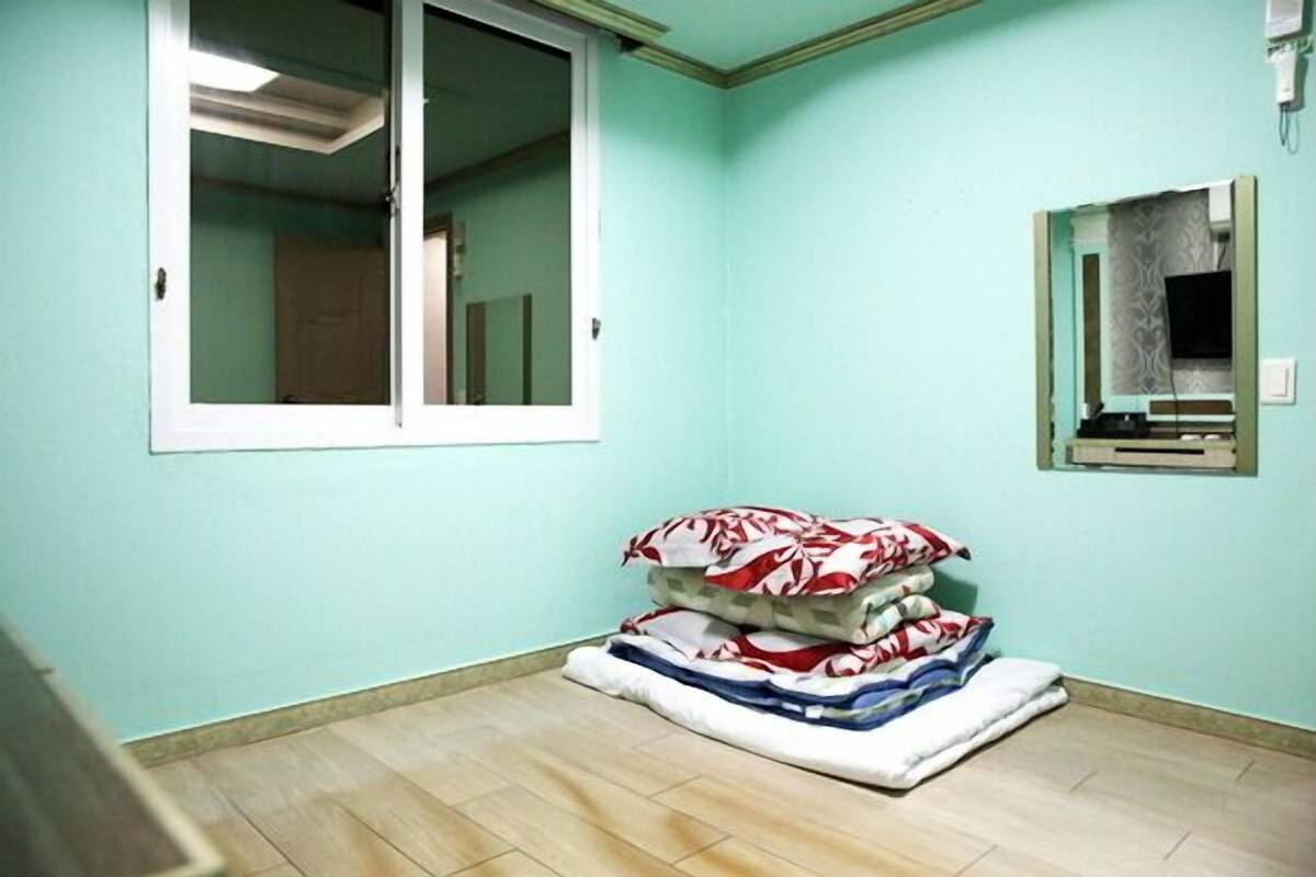 Een kamer met blauwgroene wanden, een raam met witte lijst, een spiegel en een stapel kussens op de vloer.