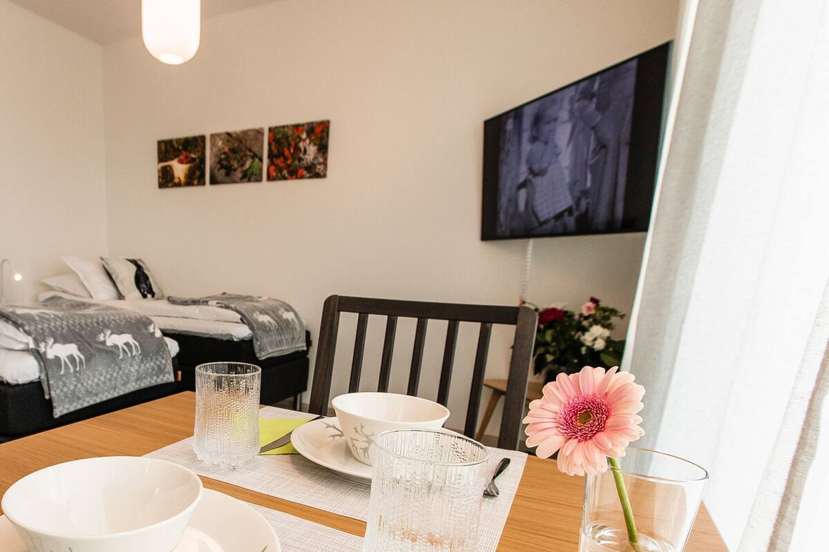 A neatly arranged room with a bed, a dining table with a flower vase, and a TV mounted on the wall.
