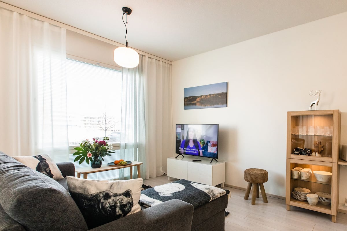 A modern living room with a grey sofa, a TV on a white stand, a wooden sideboard, and a window with sheer curtains.