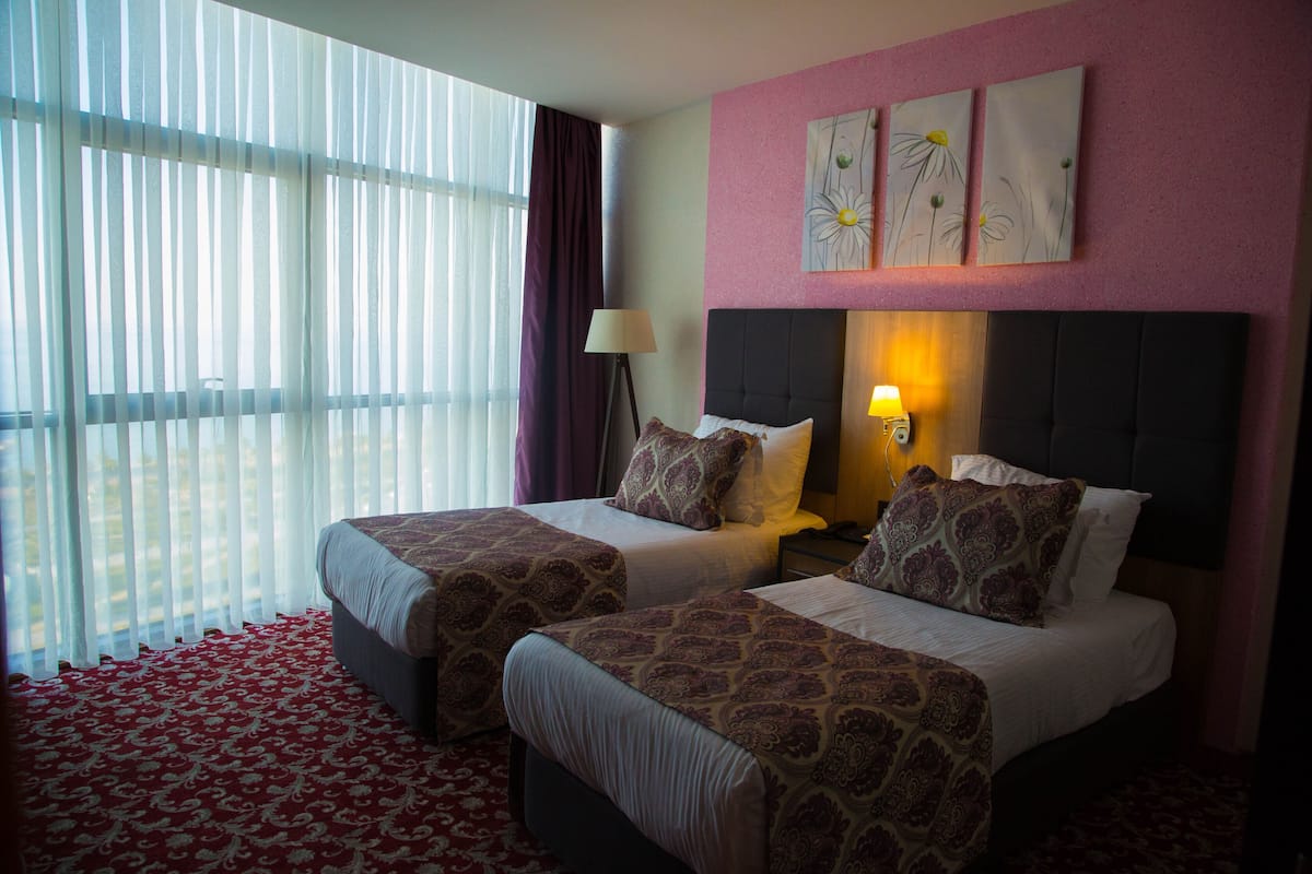 Habitación de hotel con dos camas, un ventanal con cortinas translúcidas, una lámpara de pared y dos cuadros con flores.