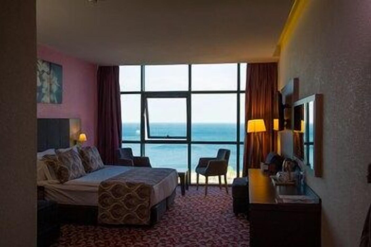 A hotel room with a bed, a desk, a chair, and a large window with a view of the sea.