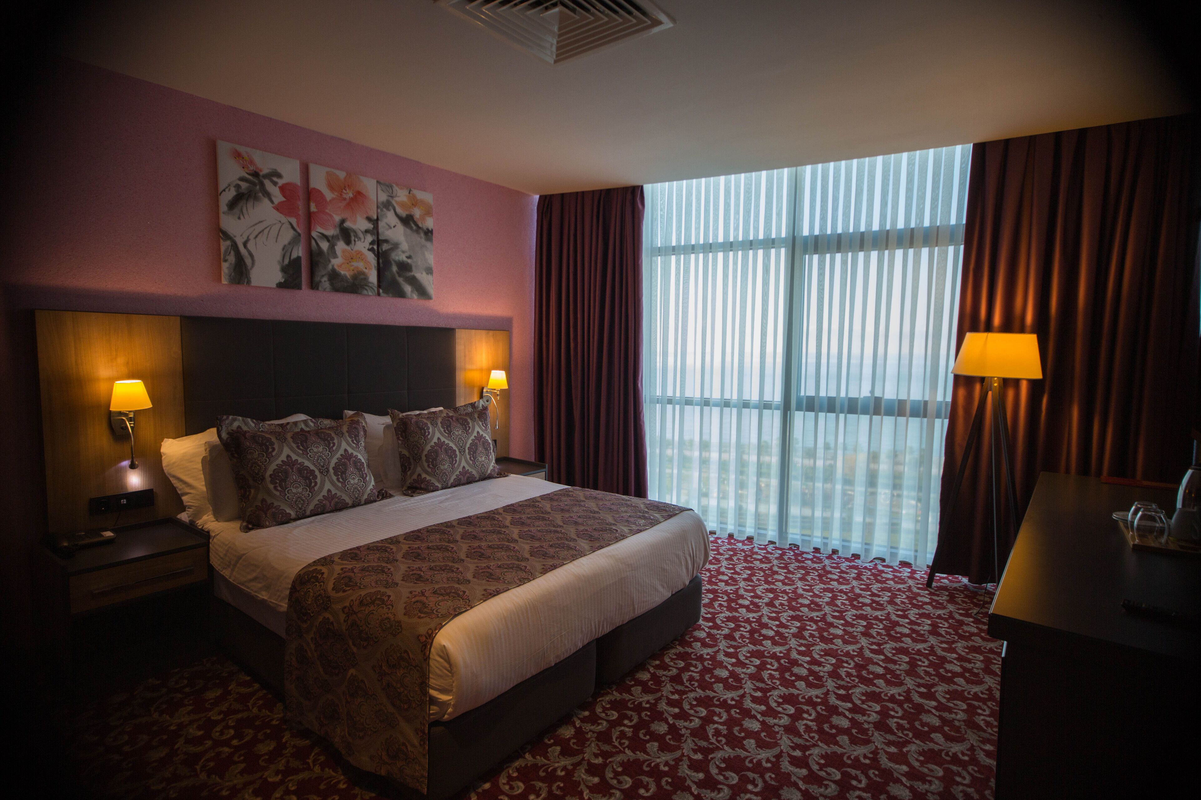 Een hotelkamer met een groot bed, twee bedlampen, een hoofdeinde, een raam met gordijnen, en een wand met bloemwerk.