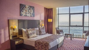 Habitación de hotel con una cama grande, dos mesitas de noche con lámparas, una silla, una mesita pequeña y un ventanal con vista al mar.