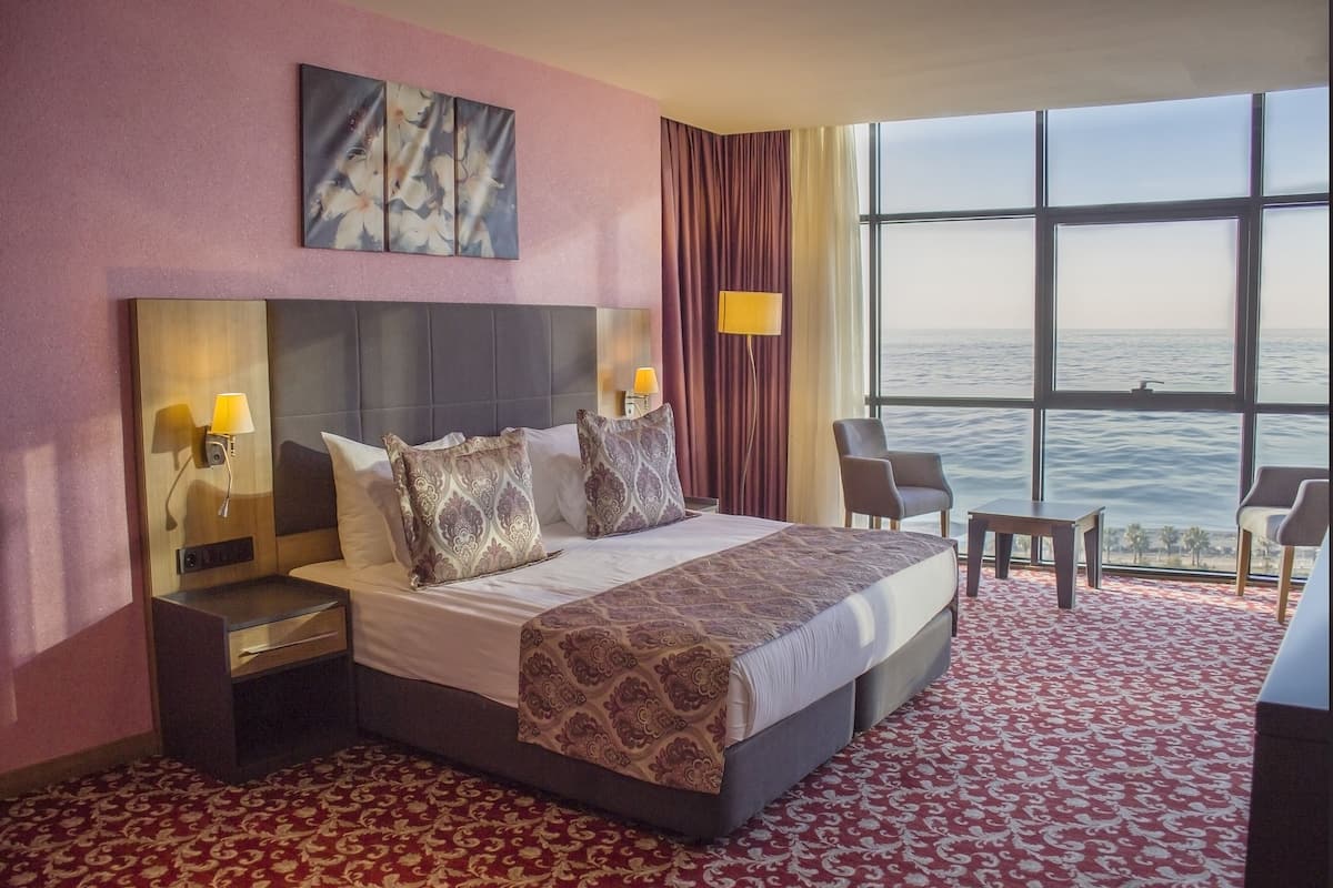 Habitación de hotel con una cama grande, dos mesitas de noche con lámparas, una silla, una mesita pequeña y un ventanal con vista al mar.