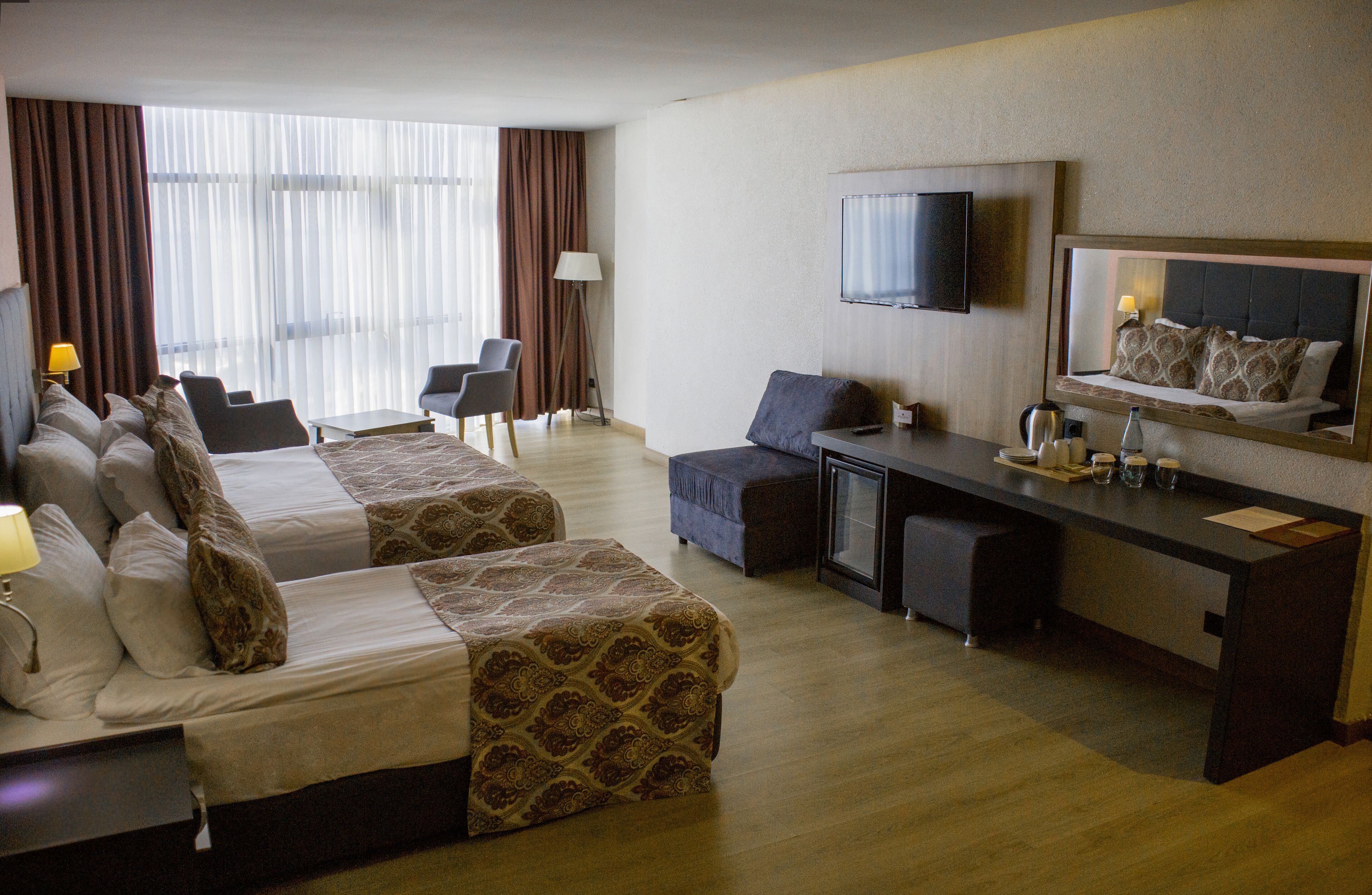 A hotel room with two beds, a desk, a chair, a TV, and a large window with curtains.