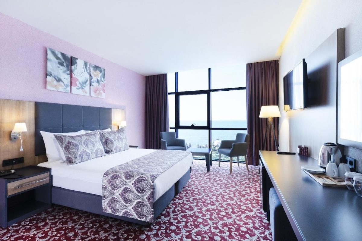 A hotel room with a bed, bedside tables, a desk, a chair, a TV, and a large window with a view.