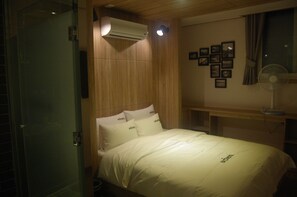 A hotel room with a bed, an air conditioning unit, a bedside table, a fan, and a wall with framed pictures.