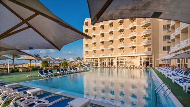 Een zwembad met ligbedden en parasols, omringd door een meerlaags hotelgebouw.