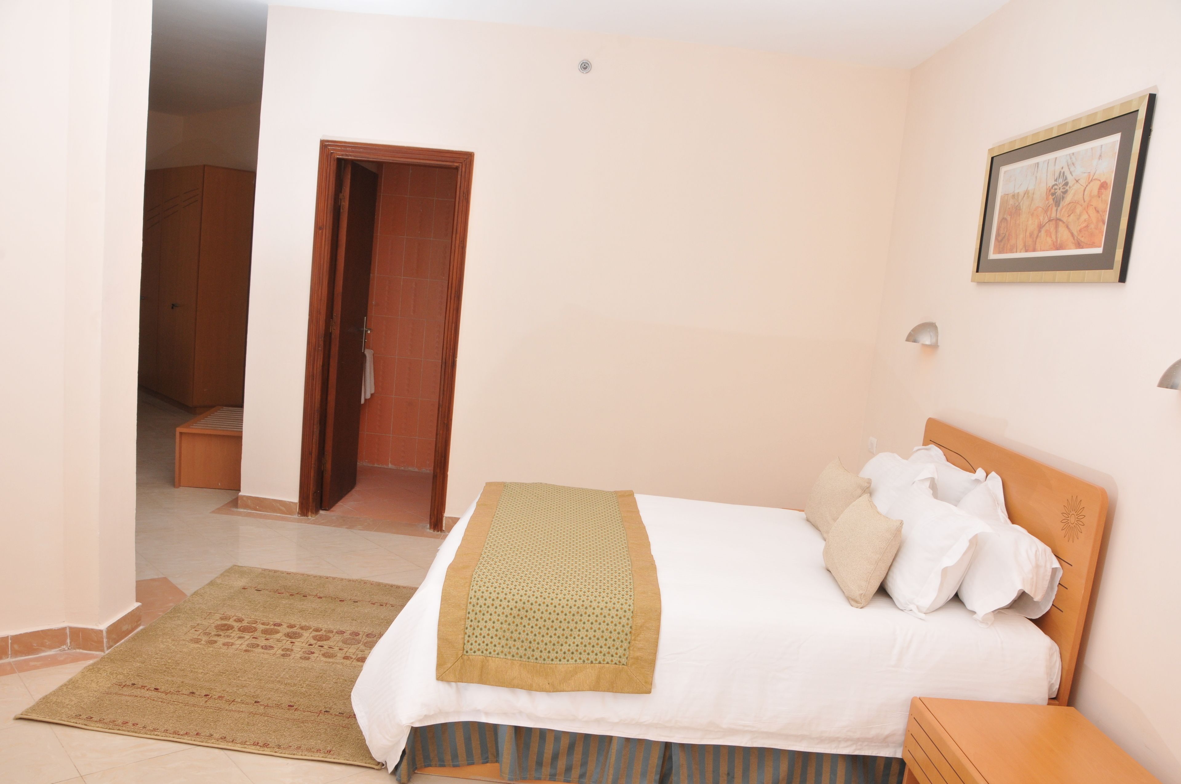 Un dormitorio con cama, almohadas, mesita de noche, cabecera de madera, lámpara de pared, puerta y pasillo.