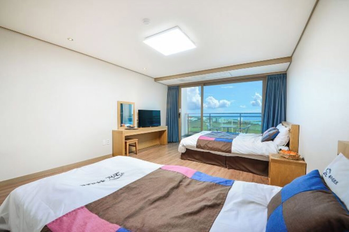 A hotel room with two beds, a desk, a TV, and a large window with a view of the sea.
