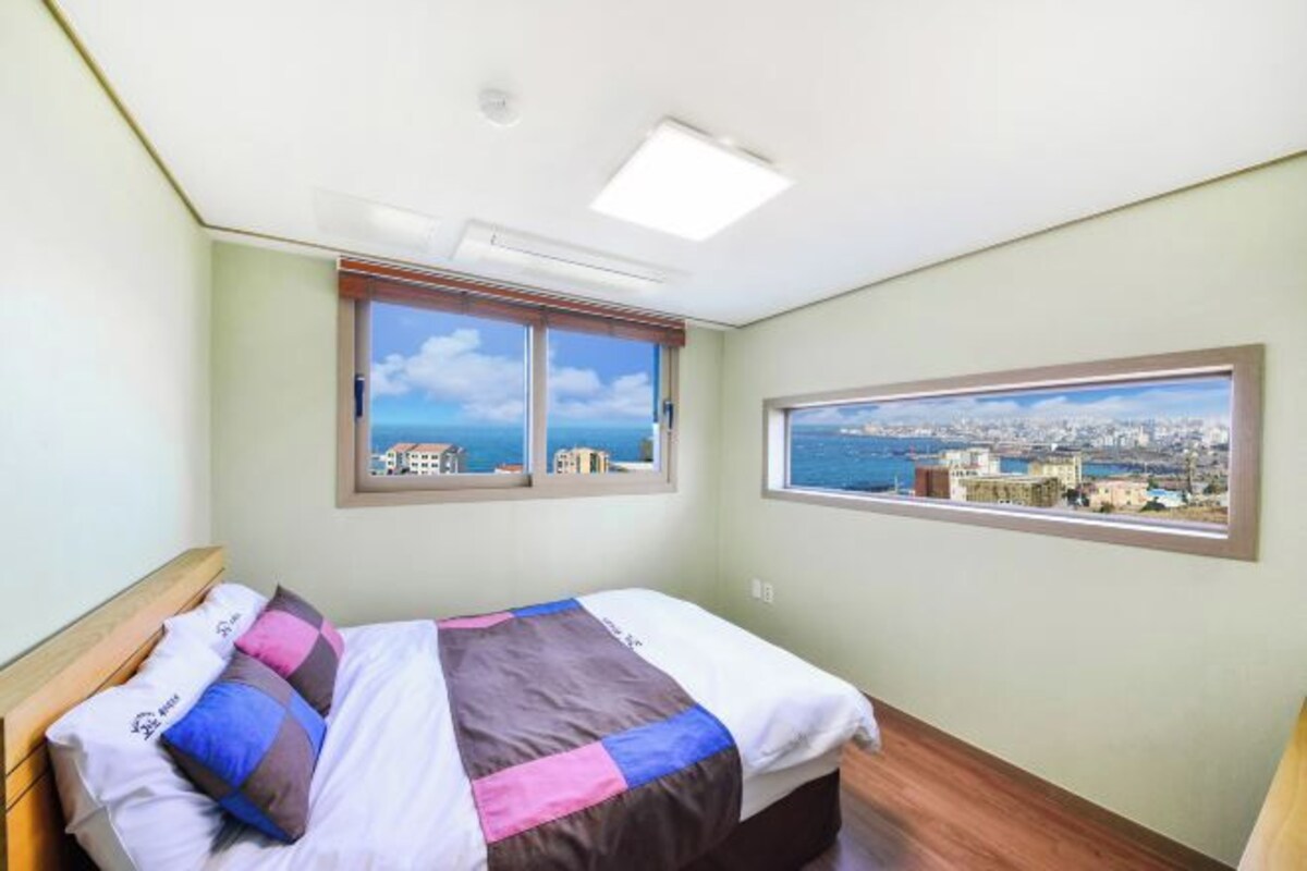 A bedroom with a bed, two windows, and a city view.