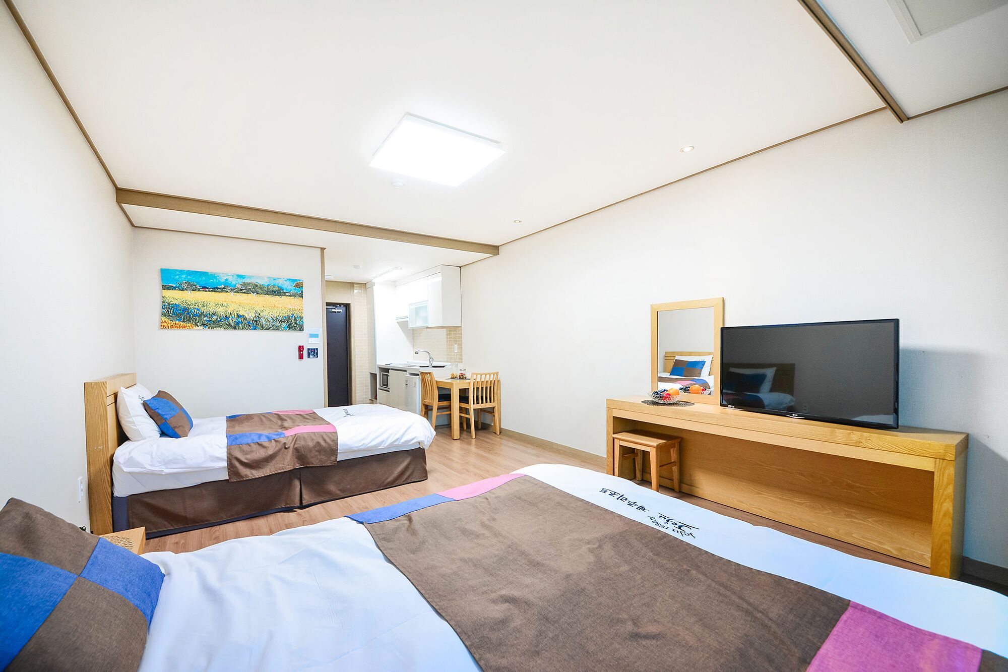 A hotel room with two beds, a desk with a TV, a small kitchenette, and a painting on the wall.