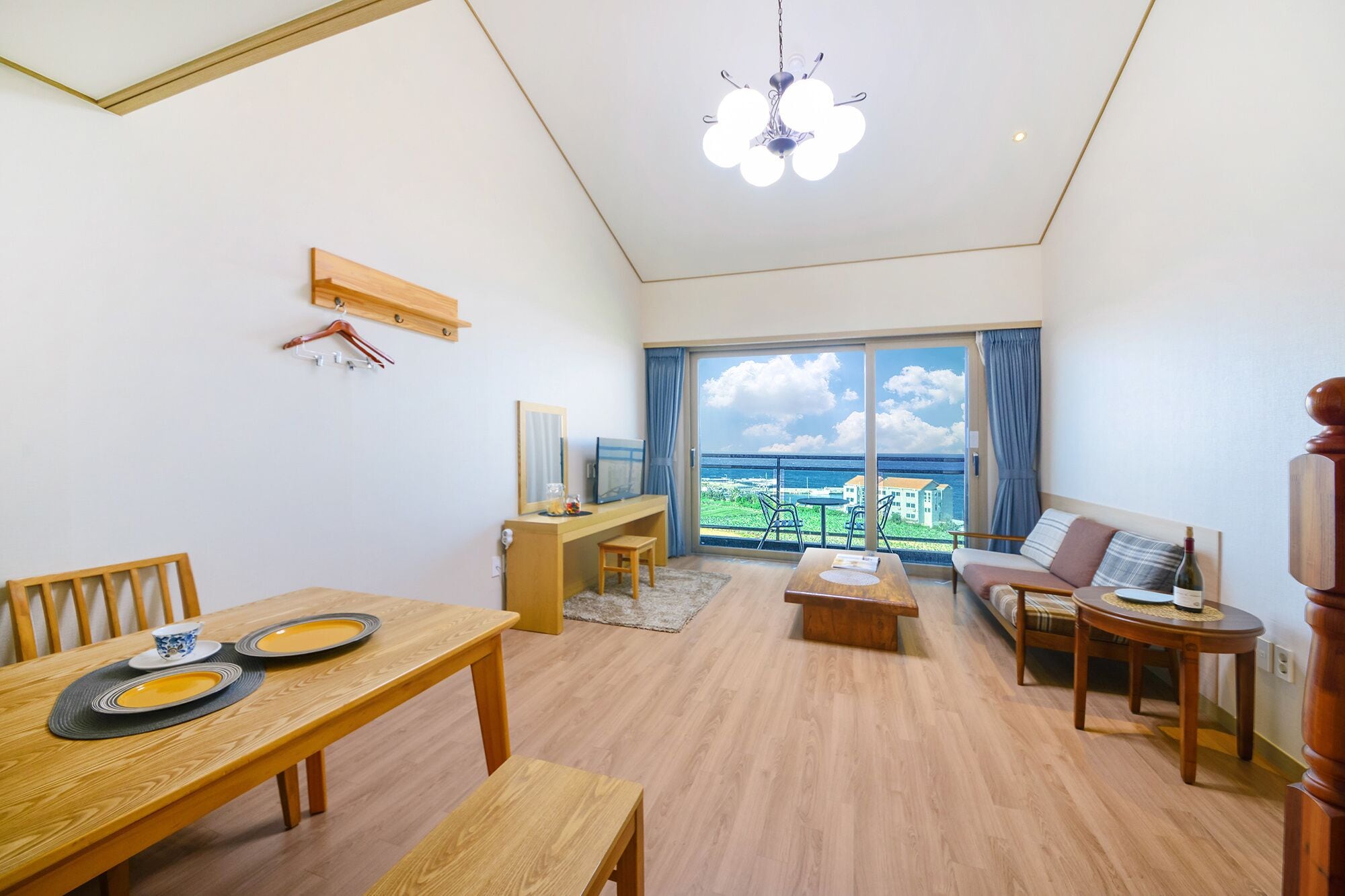 A spacious living room with a wooden floor, a dining table with chairs, a sofa, a coffee table, and a balcony with a view of the sea.
