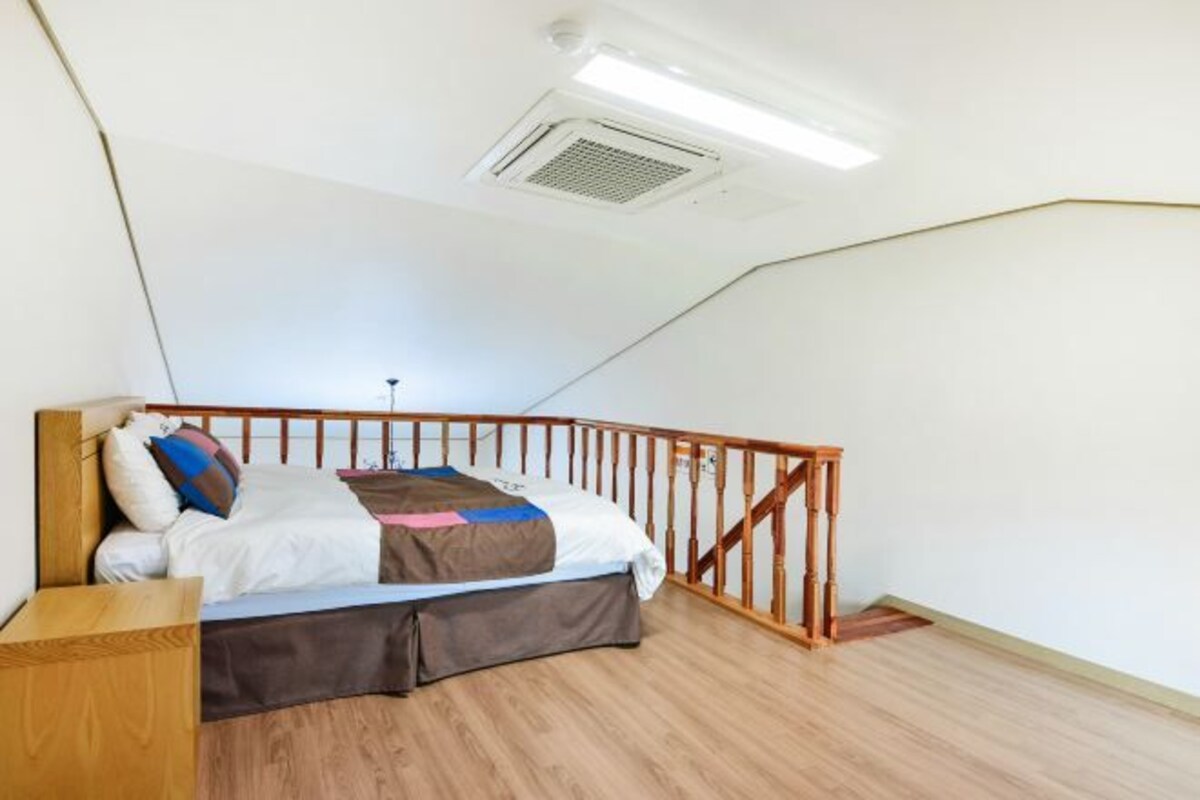 A bedroom with a sloped ceiling, a wooden bed frame, a headboard, a bedside table, and a small wooden stool.