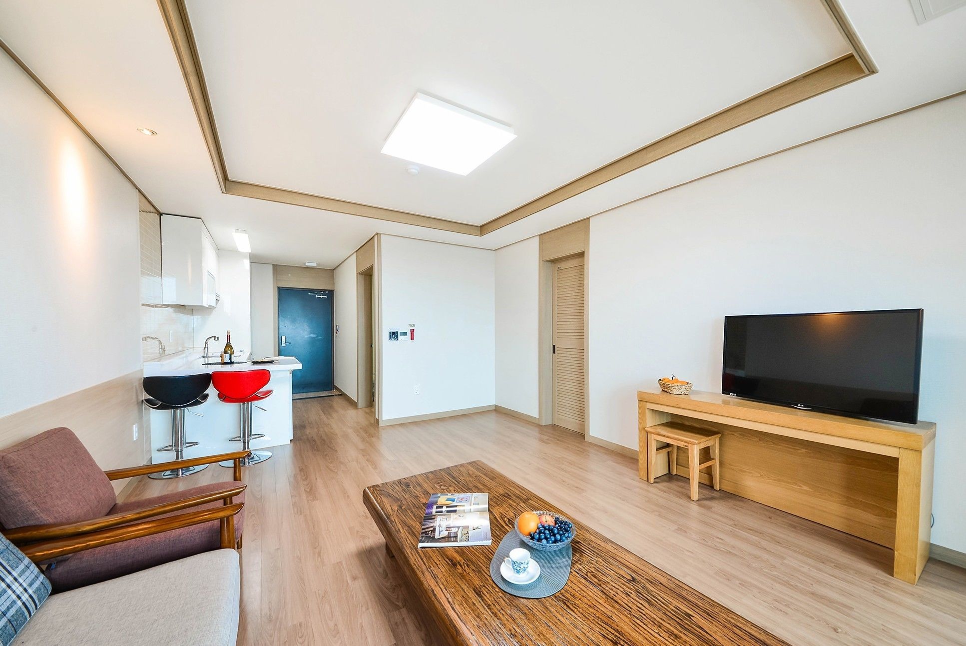 A modern living room with a wooden coffee table, a TV on a wooden stand, a red chair, and a kitchen area with a blue door.