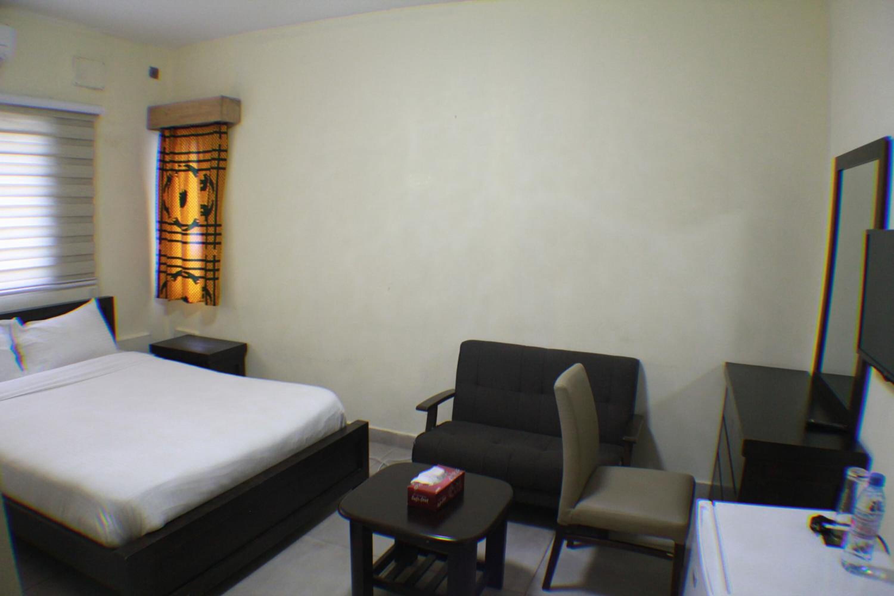 Quarto de hotel com cama, uma pequena mesa com uma caixa, uma cadeira, escrivaninha com computador e televisão.