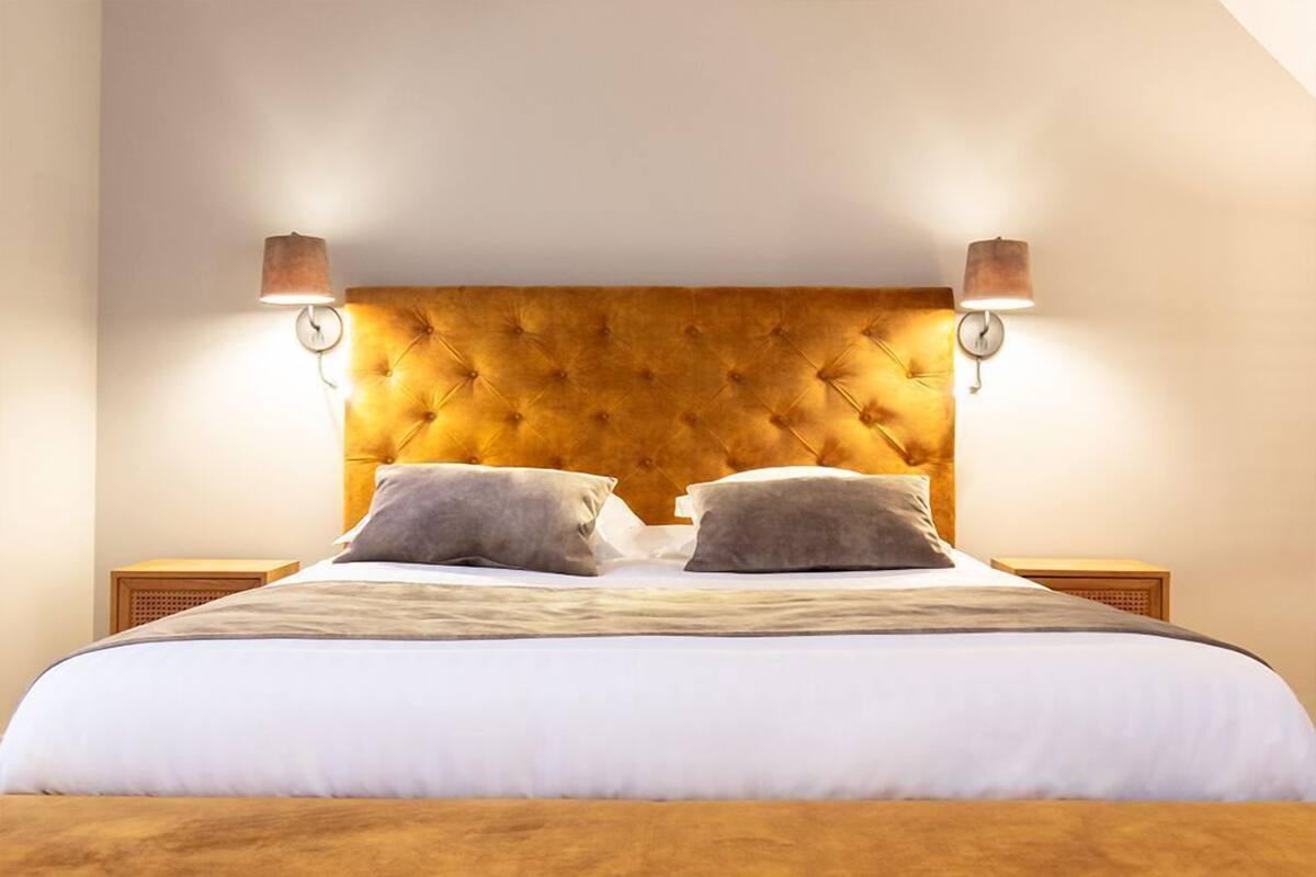 Cama bem arrumada com cabeceira estofada, duas luminárias de apoio e esquema de cores neutras.