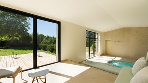Een modern interieur met een grote schuifdeur naar een houten terras, een kleine tafel, een comfortabele bank en een wellnessruimte met een jacuzzi.