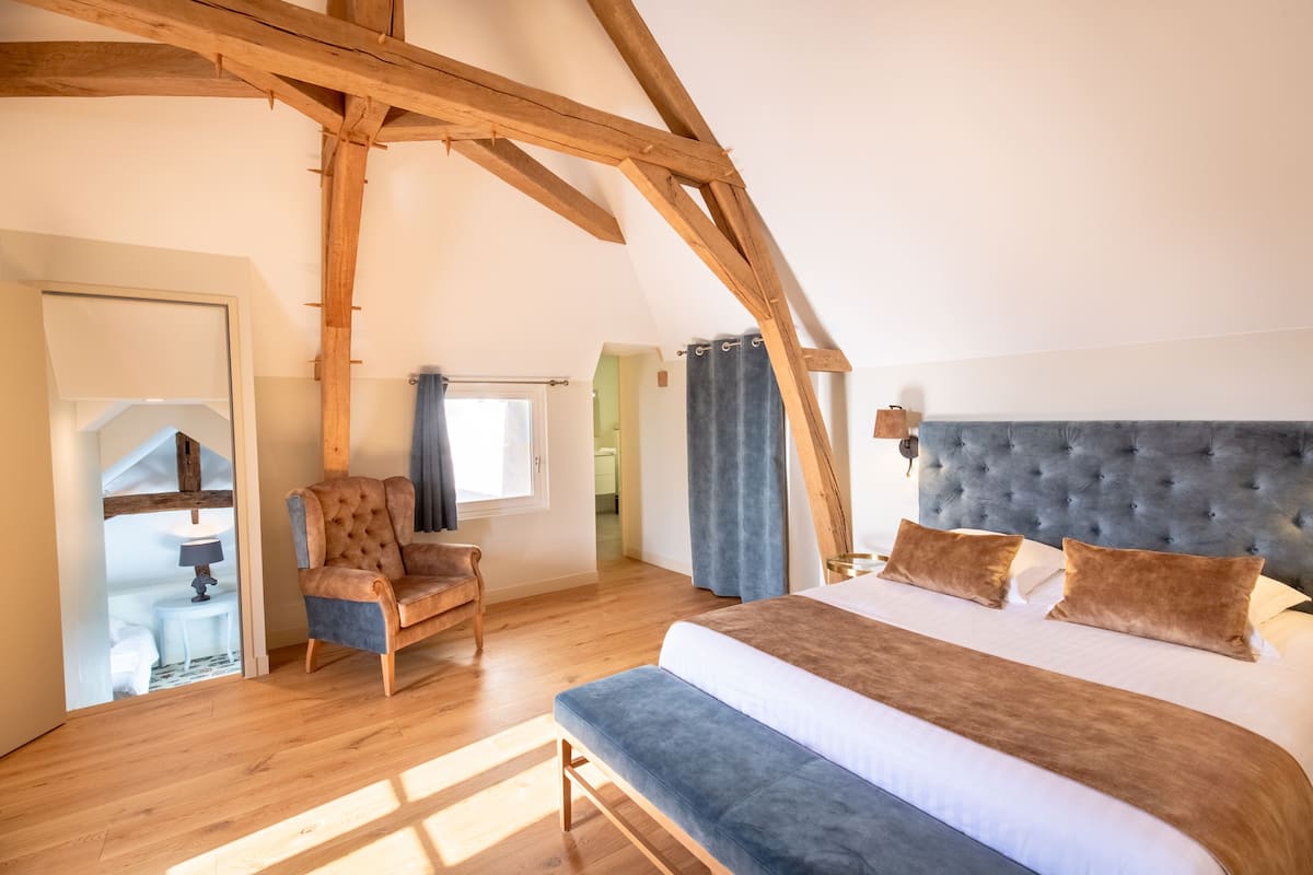 Quarto com cama grande, uma cadeira, vigas de madeira, janela e porta.