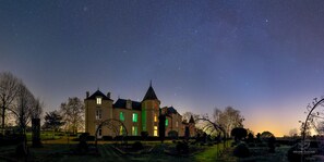 Un edificio grande e histórico con múltiples torres y ventanas iluminadas en verde, rodeado de un jardín cuidado y árboles.